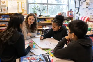 Small group of students and a teacher collaborating at a busy table with shared resources in a naturally lit, lived-in classroom.