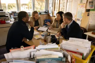 Teachers of different ages working together in a lively, sunlit staff room, intensely discussing and developing innovative cooperative teaching strategies around a table scattered with materials and digital tools.