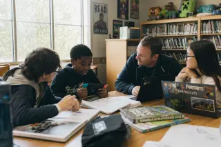Small group of middle school students and a teacher collaborating around a table with game strategy notes and laptops in a library, immersed in discussion with afternoon light and a relaxed, authentic atmosphere.