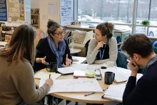 Educators in a sunlit staff lounge collaborating over lesson plans and analysis grids, showing engaged and authentic exchange.
