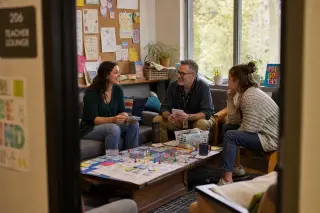 Three teachers in a sunlit break room discussing game-based learning around a table with educational games and creative materials.