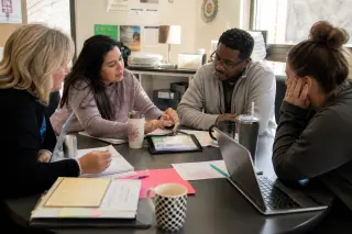 Teachers of mixed ages engaged in collaborative discussion around a table in a bright, authentic staff room, referencing a digital platform on a tablet amid notes and coffee mugs.