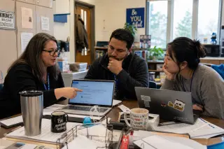 Three teachers in a relaxed staff room, discussing data privacy settings on laptops, natural light and authentic interaction.