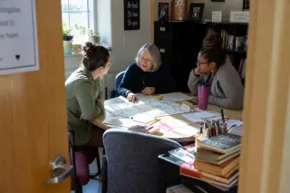 Three teachers of varying ages collaborate over a board game prototype in a sunlit, lived-in teachers' lounge filled with teaching materials, captured candidly from behind an open door.