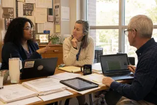 Three teachers of different ages in a sunlit, authentic staff room, deeply engaged in collaborative discussion and feedback, gathered around a table with papers, notebooks, and digital devices, in a relaxed, natural atmosphere.