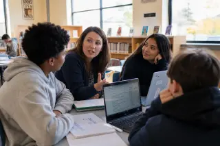 Teacher and three students in a bright library collaborating around laptops and notebooks, engaged in a discussion about educational technology use.