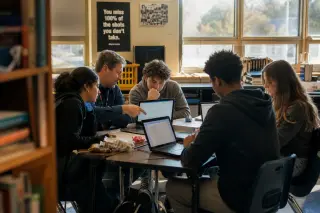 Teacher and four high school students in a relaxed classroom corner, collaborating with laptops and papers, sunlight streaming in, captured naturally from behind a shelf.