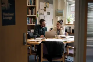 Two teachers in a relaxed staff room talk intently over a laptop, lesson materials scattered between them, afternoon light creating a warm, collegial ambiance.