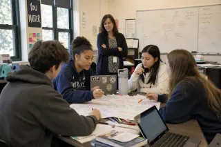 High school students collaborating around a table covered with notes and laptops in a lively, authentic classroom while a teacher observes in the background
